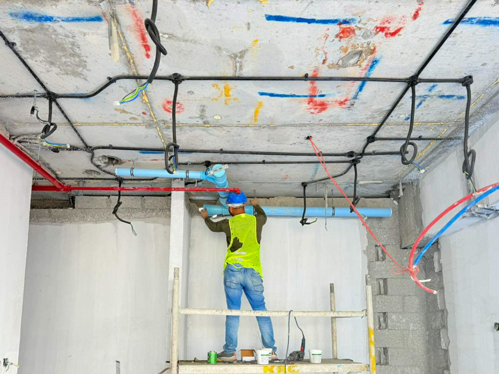 Beach Walk Iii By Imtiaz A worker in a green vest is installing pipes on a ceiling while standing on a ladder in a construction area.